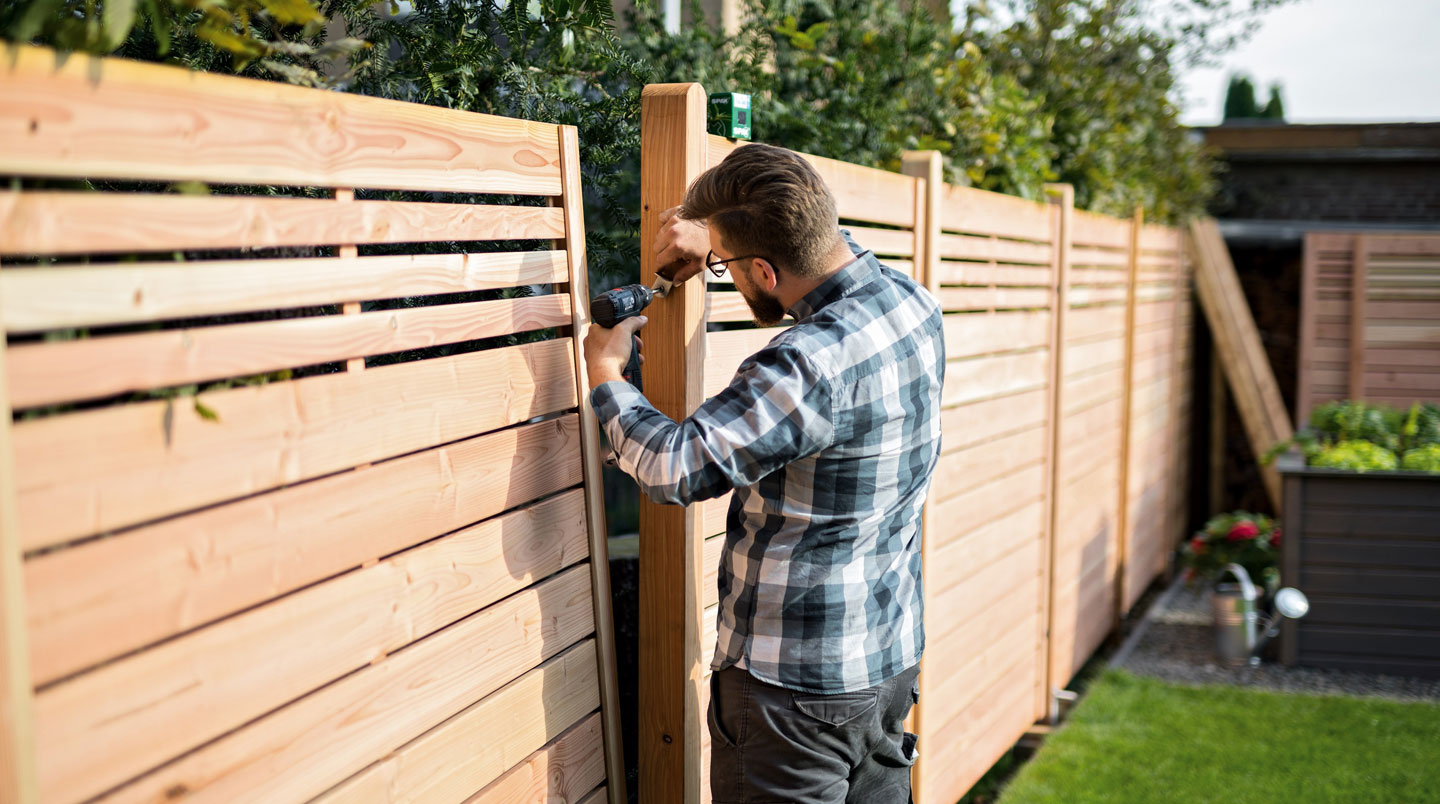 Technicien professionnel debout en tenue de chantier installant une pergola bioclimatique aluminium blanche sur terrasse composite, visseuse électrique en main, matériaux au sol, jardin résidentiel en arrière-plan