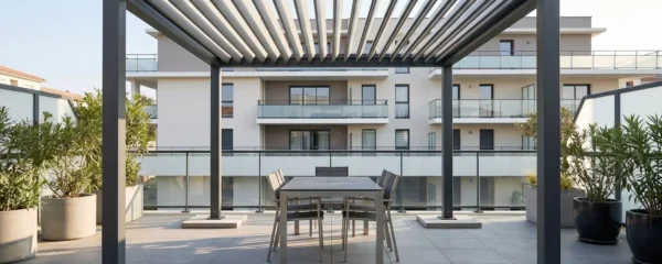 Pergola autoportée en aluminium anthracite installée sur terrasse de copropriété moderne, lames orientables semi-ouvertes, mobilier de jardin dessous, dalles de lestage visibles aux pieds de la structure